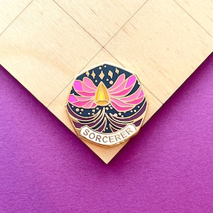May include: Gold-toned enamel pin with a black and purple background. The pin features a pink flower with a yellow centre and the word "SORCERER" in a white banner.