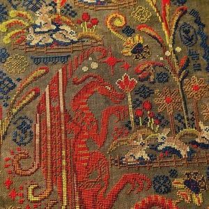 May include: Framed cross-stitch artwork featuring a detailed, colorful design. The piece showcases a central red figure with intricate patterns, surrounded by floral and foliate motifs in shades of blue, yellow, and brown. The artwork is encased in a wooden frame.