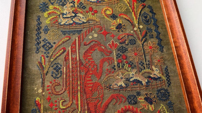 May include: Framed cross-stitch artwork featuring a detailed, colorful design. The piece showcases a central red figure with intricate patterns, surrounded by floral and foliate motifs in shades of blue, yellow, and brown. The artwork is encased in a wooden frame.