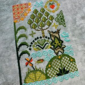 K&ouml;nnte beinhalten: Kreuzstich-Stickerei, die eine fantasievolle Szene mit einem Baum, einer Blume, einem Vogel und einem kleinen Wesen zeigt. Das Design ist in Gr&uuml;n-, Gelb-, Orange- und Blaut&ouml;nen auf einem wei&szlig;en Stoff gestickt.