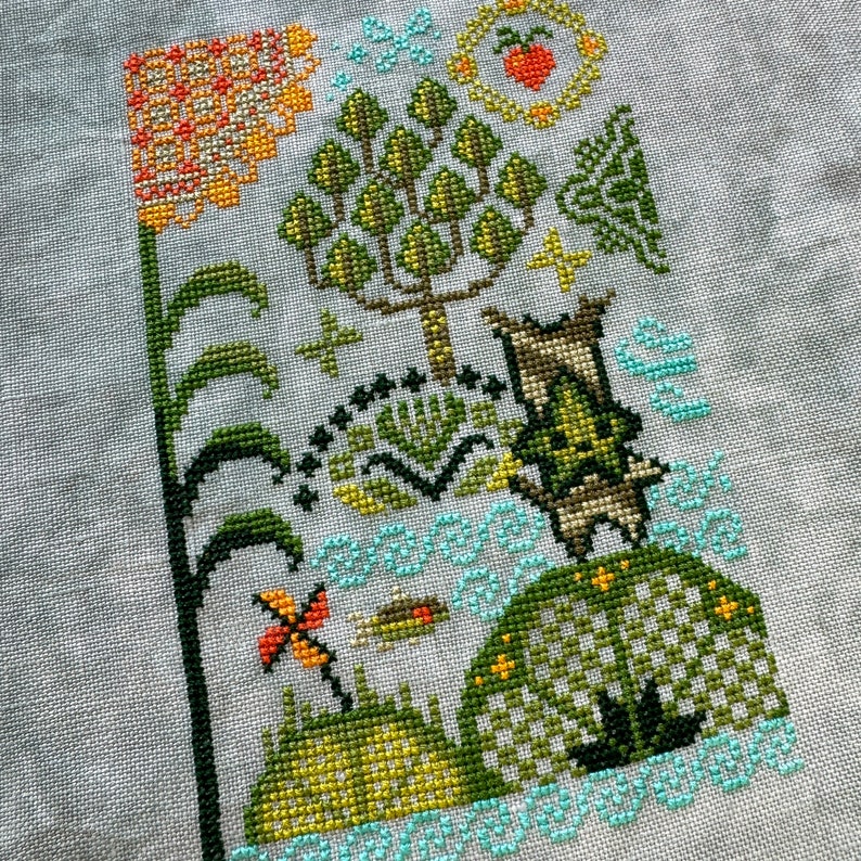 K&ouml;nnte beinhalten: Kreuzstich-Stickerei, die eine fantasievolle Szene mit einem Baum, einer Blume, einem Vogel und einem kleinen Wesen zeigt. Das Design ist in Gr&uuml;n-, Gelb-, Orange- und Blaut&ouml;nen auf einem wei&szlig;en Stoff gestickt.