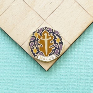 May include: A gold-colored enamel pin with a sword design, surrounded by purple and yellow accents. The word "MAGUS" is written in white at the bottom. The pin is set against a light wood background.