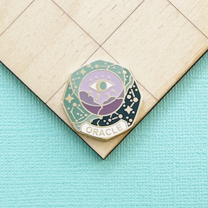 May include: A colorful enamel pin with an oracle theme. The pin features an eye design with a crescent moon, stars, and the word "ORACLE". The pin has a gold border and is set against a light wood surface.