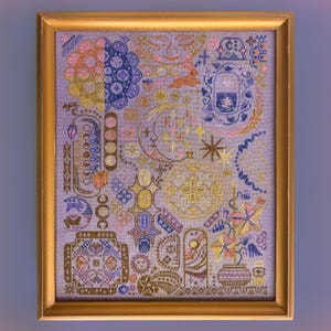 May include: A framed cross-stitch embroidery on light purple fabric featuring a collection of celestial and botanical motifs including moons, stars, flowers, and geometric patterns in shades of gold, blue, pink, and brown within a gold frame.