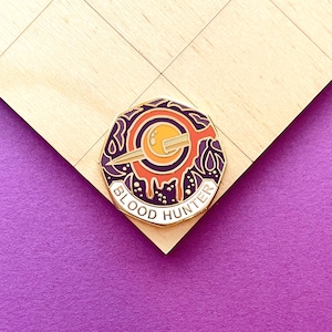 May include: A round enamel pin with a gold border, featuring a stylized design with a crescent moon, a sword, and the text "BLOOD HUNTER". The pin has a color scheme of orange, purple, and white.