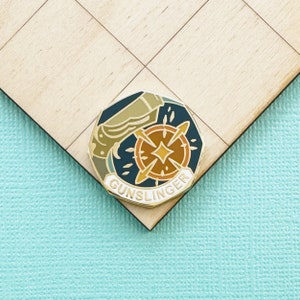 May include: A gold-colored enamel pin with a blue, brown, and orange design. The pin features a stylized compass with a gun and the word "GUNSLINGER" around the edge.