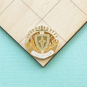 May include: A Champion enamel pin with a shield and sword design, featuring a cross in the center. The pin has a gold border and is set against a light wood surface and a turquoise background.
