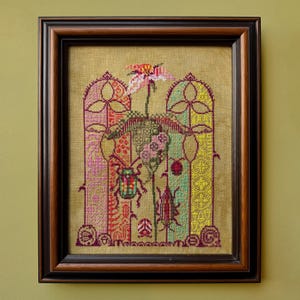 May include: Framed cross-stitch artwork featuring a central flower and insects. The design includes colorful vertical panels with floral and geometric patterns. The frame is dark brown, and the background is a light tan color.