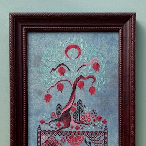 Puede incluir: Un bordado de punto de cruz enmarcado que representa un &aacute;rbol estilizado con frutos rojos y una luna creciente. El &aacute;rbol est&aacute; rodeado por un borde de flores y hojas rojas m&aacute;s peque&ntilde;as. El fondo es una tela azul claro.