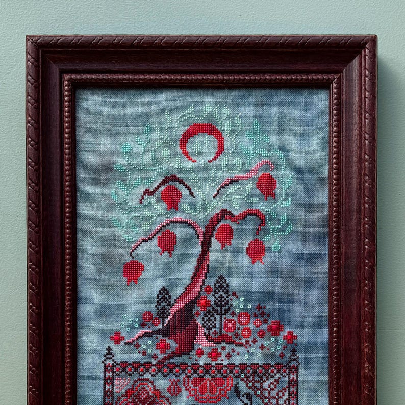 Puede incluir: Un bordado de punto de cruz enmarcado que representa un &aacute;rbol estilizado con frutos rojos y una luna creciente. El &aacute;rbol est&aacute; rodeado por un borde de flores y hojas rojas m&aacute;s peque&ntilde;as. El fondo es una tela azul claro.
