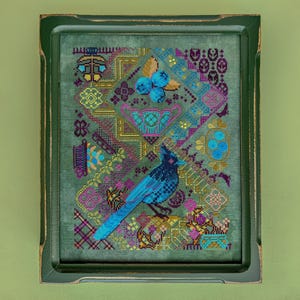 以下が含まれることがあります： 額縁に入ったクロスステッチアート。長い尾を持つ青い鳥が、花柄のデザインの上に止まっています。作品は、ティール、紫、金色の幾何学模様と花柄のモチーフが施された背景に設定されています。フレームは緑色です。