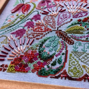 May include: A cross-stitch embroidery piece on light blue fabric featuring a butterfly motif. The design incorporates intricate floral patterns in shades of pink, red, green, and brown, accented with metallic bugle beads and small seed beads.