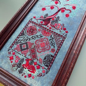 Puede incluir: Un bordado de punto de cruz enmarcado que presenta un &aacute;rbol estilizado con frutos rojos y una luna creciente. El &aacute;rbol est&aacute; rodeado de un patr&oacute;n floral en tonos rojos, negros y blancos sobre un fondo azul.