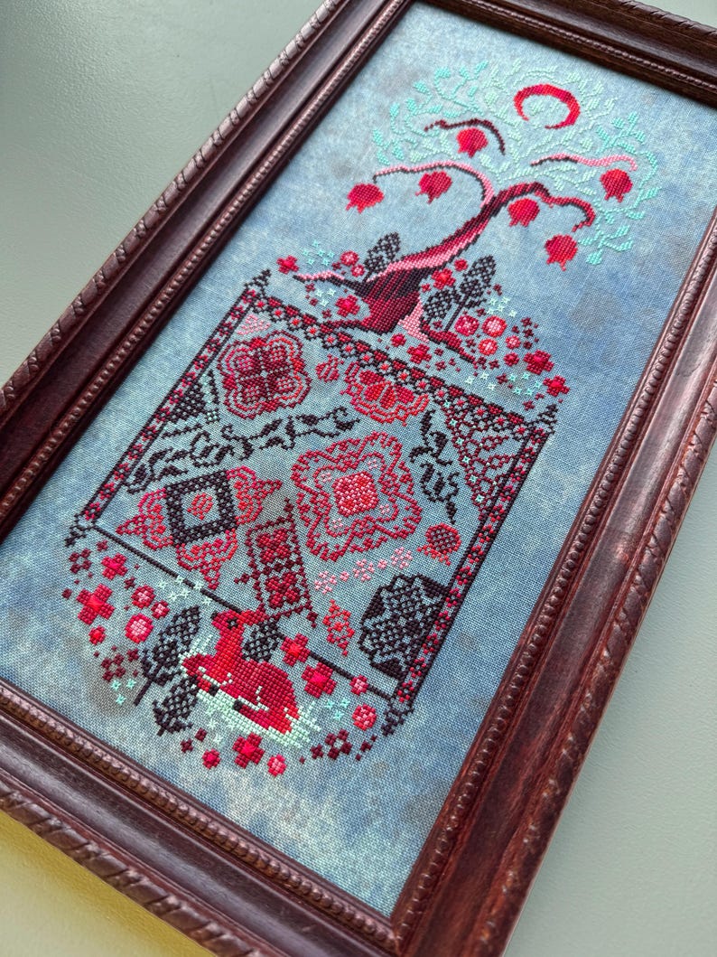 Puede incluir: Un bordado de punto de cruz enmarcado que presenta un &aacute;rbol estilizado con frutos rojos y una luna creciente. El &aacute;rbol est&aacute; rodeado de un patr&oacute;n floral en tonos rojos, negros y blancos sobre un fondo azul.