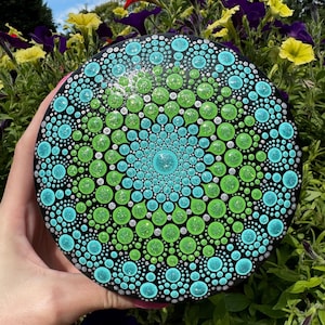 Puede incluir: Una piedra redonda, pintada de negro, con un diseño de mandala en tonos de verde, azul y plateado. El diseño está formado por pequeños puntos y círculos.
