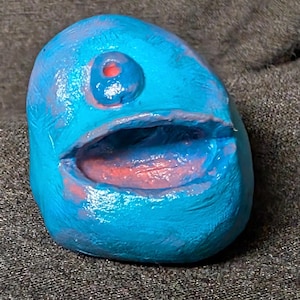 May include: A blue, abstract sculpture with a single red eye and an open mouth revealing a red interior. The artwork has a textured surface and is set against a gray background.