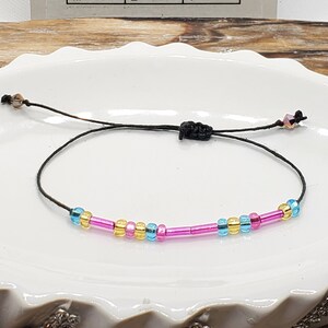 May include: A black string bracelet with pink, blue, and gold beads. The bracelet is on a white plate. The Morse code alphabet is visible in the background.