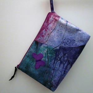 May include: A purple and blue fabric pouch with a zipper closure and a purple butterfly applique. The pouch has a watercolor-like design with abstract shapes and lines.