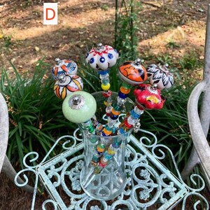 May include: A glass vase filled with colorful beaded sticks topped with ceramic knobs. The knobs are painted with various patterns and colors, including white, blue, red, orange, and green. The vase is sitting on a light blue metal table with a decorative design.