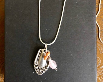 May include: A silver spoon necklace with a pink and orange bead charm. The necklace is on a black background.