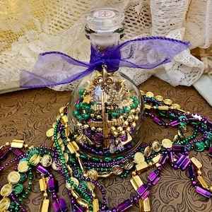 May include: A glass bottle filled with green, purple, and gold beads and charms, including a fleur-de-lis charm. The bottle is tied with a purple ribbon and surrounded by strands of green, purple, and gold beads.