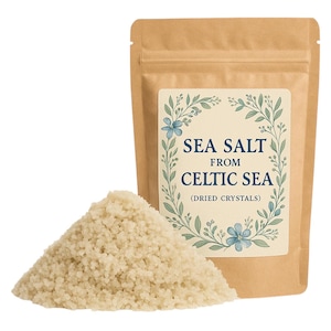 May include: A brown paper bag with a label that reads "Sea Salt from Celtic Sea (Dried Crystals)". The label has a floral design with blue flowers and green leaves. A pile of white sea salt is in front of the bag.