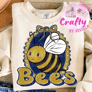 May include: Cream-colored sweatshirt featuring a cartoon bee design. The bee is yellow and black, with white wings, set against a navy blue oval frame with a gold bow. The word "Bees" is written in gold script.