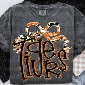 以下が含まれることがあります： オレンジと黒で「Tigers」の文字が描かれたダークグレーのスウェットシャツ。ギフトのようにデザインされています。上部のリボンはオレンジ、黒、白の模様です。スウェットシャツはComfort Colorsブランドです。