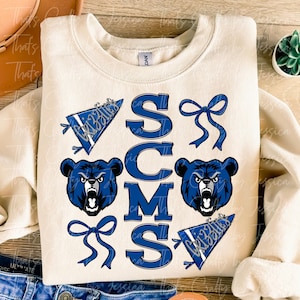 May include: A cream-colored sweatshirt with a blue and white design. The design features the letters "SCMS" in a large font, with a bear head and a pennant with the word "Grizzlies" on each side. There are also blue bows on the design.
