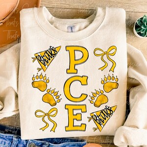 May include: Cream-colored sweatshirt with the letters "PCE" in yellow, surrounded by yellow bear paw prints, bows, and pennants that say "Bears". The sweatshirt is folded.