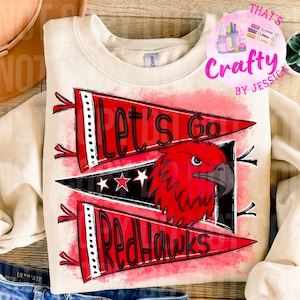 May include: Cream-colored sweatshirt with a red and black graphic. The graphic features a red hawk head and the words "Let's Go RedHawks" on pennant-shaped banners. The design includes stars and a distressed, watercolor-like background.
