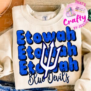 May include: Cream-colored sweatshirt with the word "Etowah" in blue bubble letters, stacked vertically, with a white trident and the words "Blue Devils" in black script. A pink circular logo is in the upper right corner.
