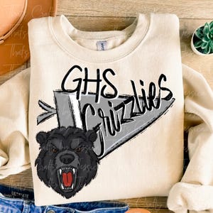 May include: Cream-colored sweatshirt featuring the text "GHS Grizzlies" in black and white, with a graphic of a snarling black bear. The design is on a gray pennant shape.