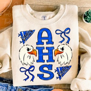 May include: Cream-colored sweatshirt with a blue and white graphic design. The design includes the letters AHS in blue, two eagle heads, and two pennants that say "Eagles". Two blue bows are also included in the design.