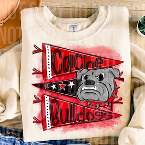 以下が含まれることがあります： 「Cache Bulldogs」の文字が入った赤と黒のペナントデザインのクリーム色のスウェットシャツ。デザインには、グレーのブルドッグのグラフィックと白い星が含まれています。スウェットシャツは柔らかい素材で作られています。