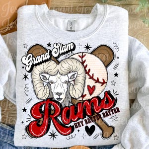 May include: A light gray sweatshirt featuring a baseball-themed graphic. The design includes a ram's head, a baseball, and a bat, with the words "Grand Slam" and "Rams" in a bold, red font. The phrase "Hey Batter Batter" is also included.
