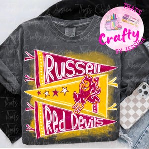 May include: A gray t-shirt featuring a design with the words "Russell Red Devils" in white, set against a yellow and maroon pennant design. The design includes a devil mascot and stars. The shirt has a vintage, distressed look.