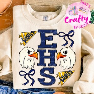 Puede incluir: Sudadera crema con un diseño gráfico azul marino "EHS", cabezas de águila y banderines "Eagles". Un logotipo rosa y morado "Crafty by Jessica" está en la esquina superior derecha. El diseño incluye lazos azul marino.