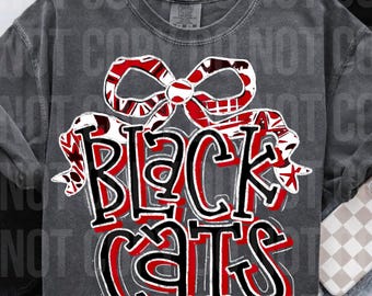 Op de afbeelding: Donkergrijze t-shirt met de tekst "BLACK CATS" in grote, vetgedrukte letters. De tekst is zwart met rode accenten en een decoratieve strik. Het shirt heeft een ronde hals.