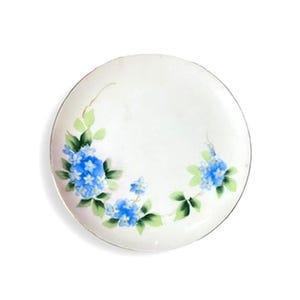 May include: White ceramic plate with a gold rim, adorned with hand-painted blue hydrangea flowers and green leaves. The floral design is arranged in a delicate pattern around the plate's edge, creating an elegant aesthetic.