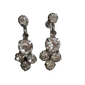 May include: A pair of vintage silver-toned clip-on earrings. Each earring features a cluster of clear, sparkling rhinestones in various sizes. The earrings have a classic, elegant design, perfect for adding a touch of glamour.