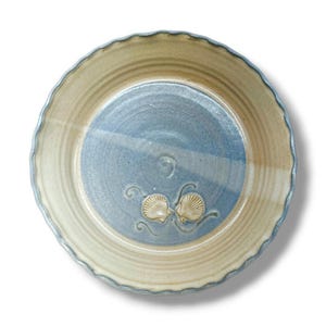 May include: A round ceramic pie dish with a scalloped edge, featuring a two-tone design with a beige outer rim and a blue center. The interior base is decorated with two raised, sculpted seashell motifs and delicate swirling line details.