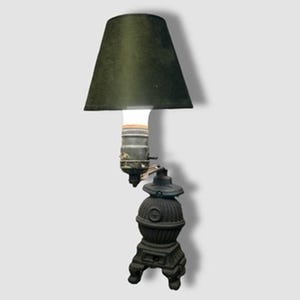May include: An antique lamp with a black lampshade and a base shaped like a miniature stove. The lamp has a vintage aesthetic, with a visible lightbulb and a dark, textured finish. The lamp is a unique decorative item.