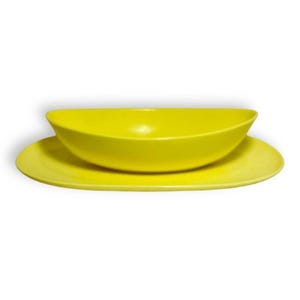 May include: A bright yellow ceramic bowl and plate set. The bowl is oval-shaped and sits atop a matching, round plate. The set has a glossy finish and is suitable for serving food. The set is a vibrant addition to any kitchen.