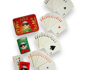 May include: A Coca-Cola branded playing card set featuring a red rectangular tin and a green box, both decorated with a vintage woman illustration. Includes four fanned sets of cards representing clubs, hearts, spades, and diamonds.