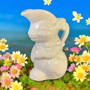 May include: A white ceramic bunny-shaped pitcher with a handle and a spout. The bunny has a bow tie and is surrounded by colorful flowers and a blue sky background. The pitcher is a decorative item.