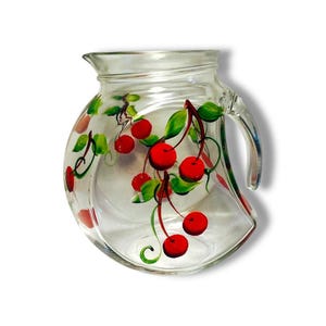 May include: A clear glass pitcher featuring a hand-painted design of bright red cherries with green leaves and stems. The pitcher has a curved, rounded body, a wide rim, and a sturdy handle on the right side, showcasing a vibrant fruit motif.