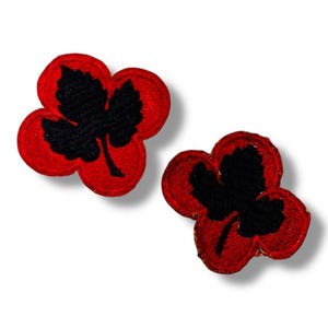 May include: Two embroidered patches, each featuring a black three-lobed leaf shape centered on a vibrant red, rounded clover-like background. The stitching is dense and textured, creating a clear contrast between the black leaf and red base.