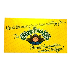 May include: A rectangular yellow card with thin diagonal lines. The text reads: Here's the news you've been waiting for... The Cabbage Patch Kids Parents' Association is about to begin! A central graphic shows a small face inside a green cabbage.