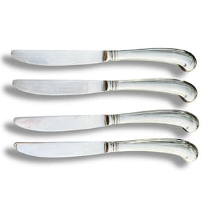May include: Four silver butter knives with curved handles and a decorative band near the blade. The knives are arranged horizontally against a white background, casting soft shadows.